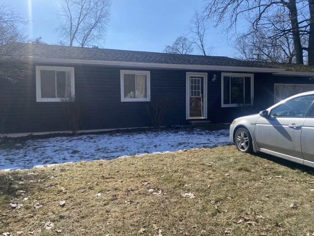 Single-story dark house with a snowy yard and a silver car parked at the right edge.