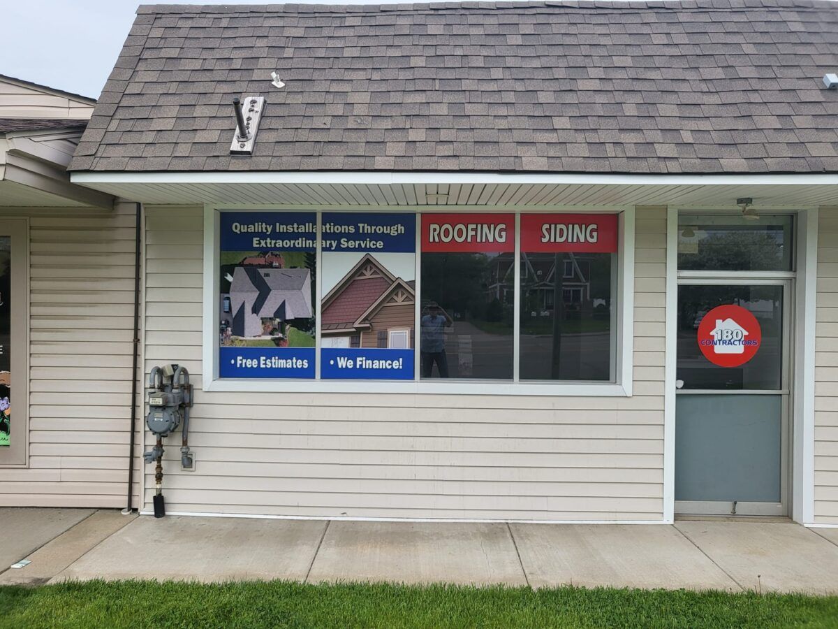 Storefront window advertising roofing and siding services, with beige siding and a glass door.