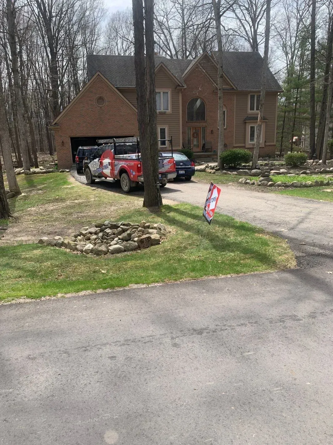 Brick house with open garage, red truck and black SUV parked in driveway, and campaign sign by the road.