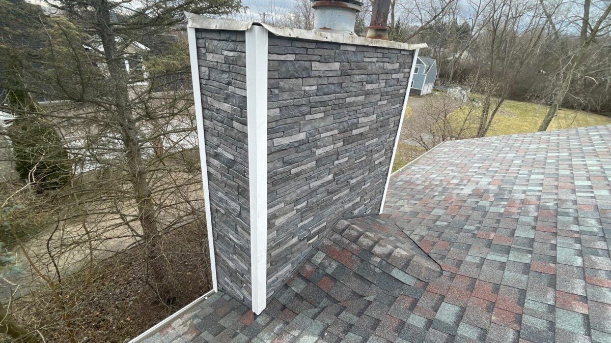 Gray stone chimney rising through a shingled roof beside bare trees under an overcast sky