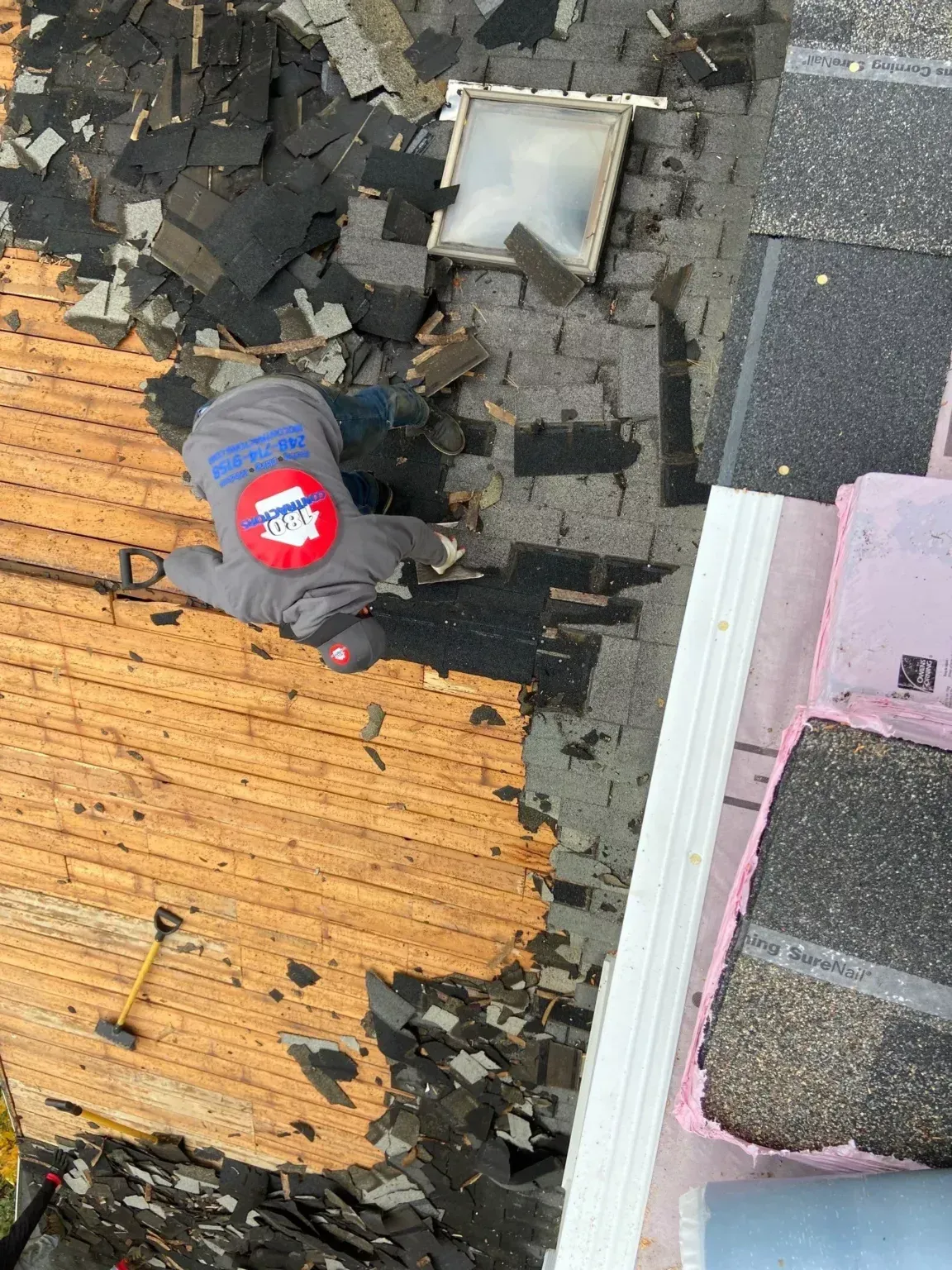 Roof worker on a partially stripped roof beside black shingles, orange sheathing, and a skylight hole