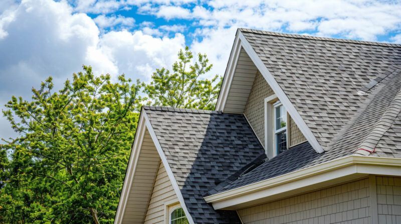 Gray shingled house roof with dormers against green trees and a blue cloudy sky