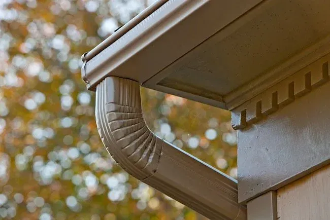 Close-up of a house gutter and downspout beneath the roof eave in autumn light