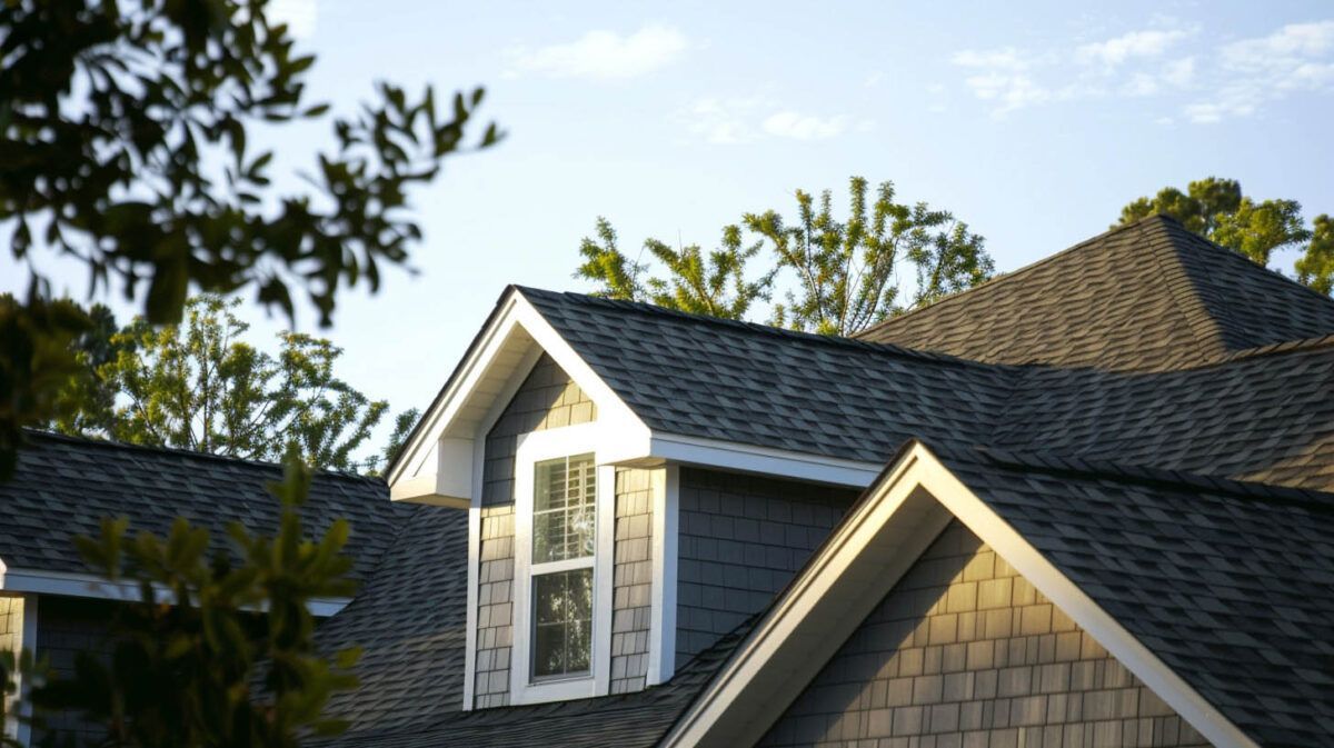 Sunlit suburban house roofs with gray shingles and dormer windows against trees and blue sky