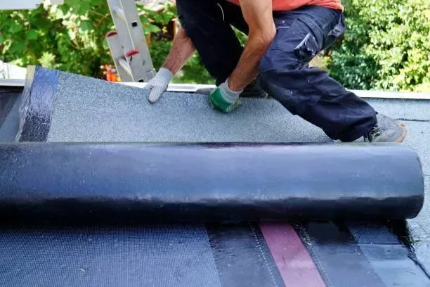 Worker installing rolled roofing on a roof, with dark gray membrane and green-gloved hands