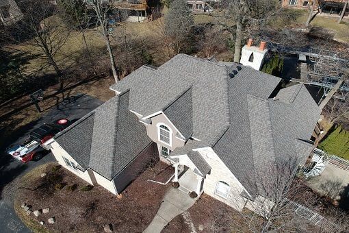 Aerial view of a gray-roofed suburban house with a driveway and leafless trees nearby.