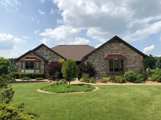 Brick house with gabled roof, landscaped front yard, and large green lawn under a partly cloudy sky