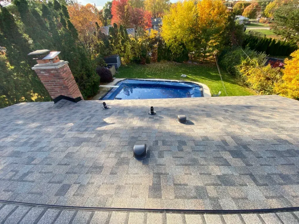 Rooftop view of a backyard with a pool, chimney, and autumn trees beyond the shingles.