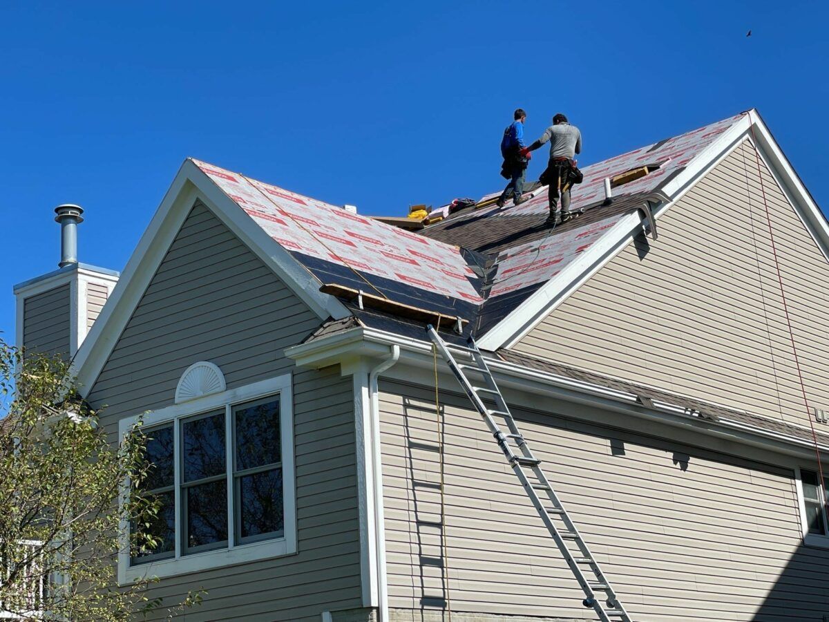 Two workers replacing shingles on a beige house roof with a ladder against the side