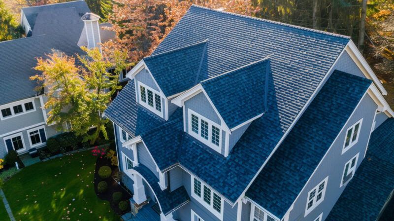 Aerial view of a large blue-gray house with dormer windows and a landscaped yard in autumn