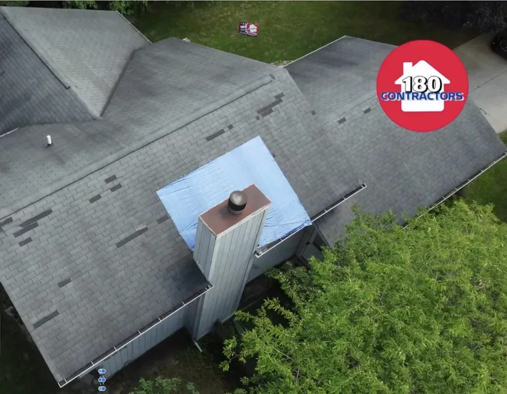 Aerial view of a gray shingled roof with a central chimney and blue roof section, surrounded by trees.