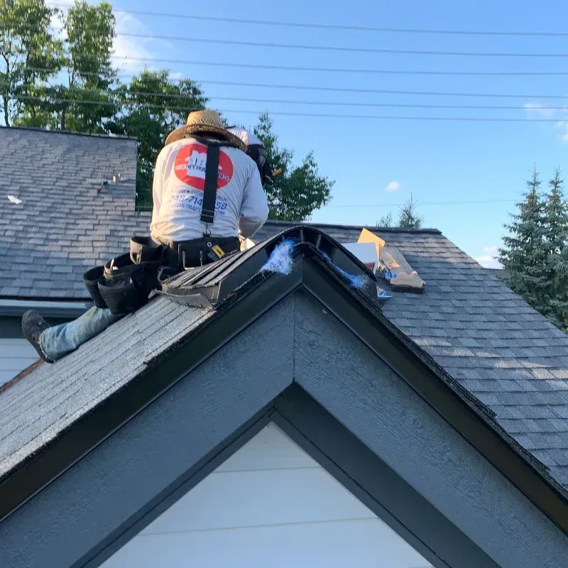 Roofing worker sitting on a house ridge, installing shingles on a gray roof.