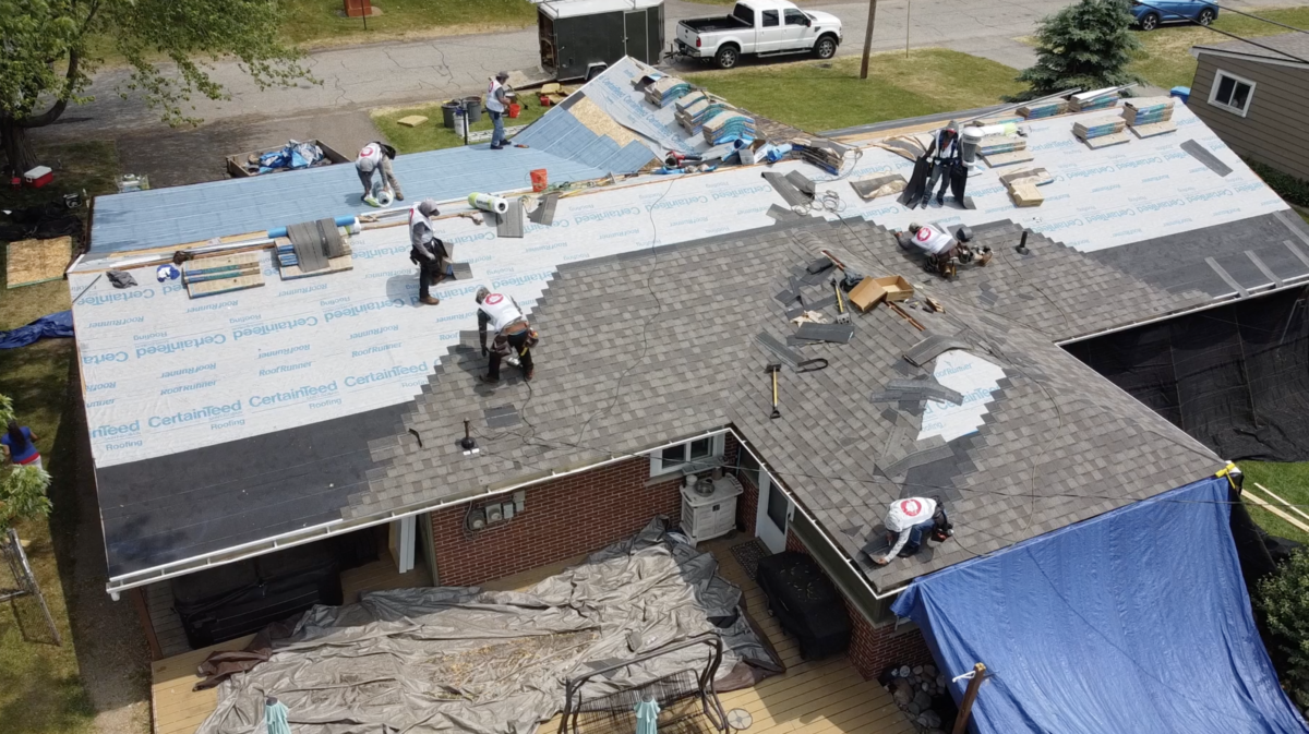 Roofing crew installing new shingles on a house roof, with materials and tools spread across sections.