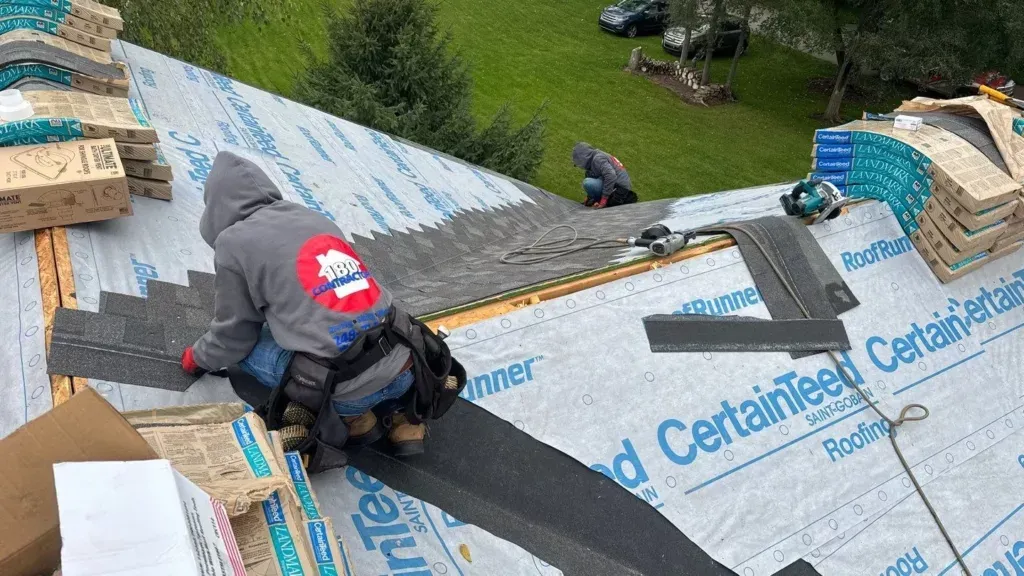 Roofing worker installing black underlayment on a steep roof near a chimney, with tools and shingles nearby.