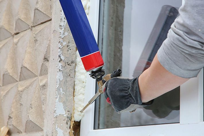 Gloved worker sealing a window frame with a caulking gun beside a stucco wall