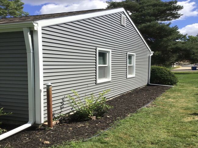 Gray house side with white-trimmed windows, downspout, and landscaped mulch bed beside a grassy yard.