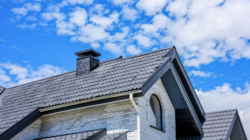 Gray shingled house roof with dormer and chimney under a bright blue sky with clouds