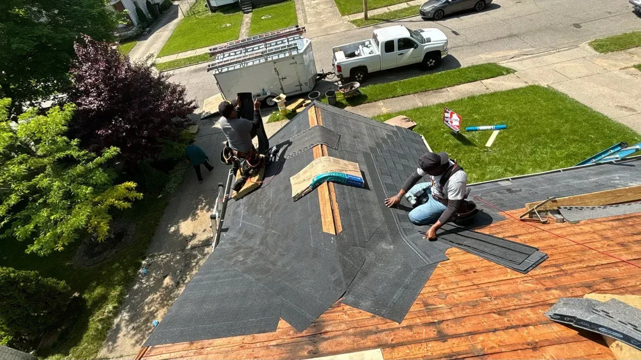 Workers installing black roofing shingles on a house roof beside a street and driveway