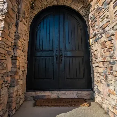 Black arched double wooden door set in a stone wall, with a small doormat at the entrance