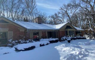 Snow-covered brick house with shrubs and bare trees in a winter yard