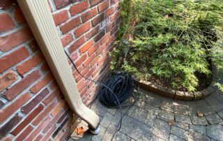 Brick house corner with downspout, coiled black hose, and paved patio beside shrubs