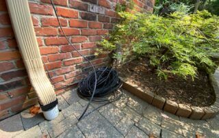 Coiled black cable by a brick wall, downspout, and garden bed on a stone patio
