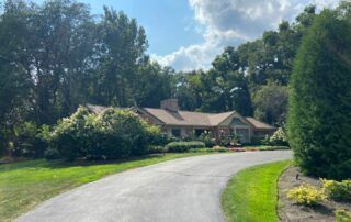 Curved driveway leading to a house surrounded by trees and manicured landscaping under a partly cloudy sky