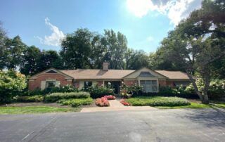 Single-story brick house with a front garden, trees, and a wide driveway under a bright blue sky
