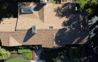 Aerial view of a tan shingle-roof house with skylights, surrounded by trees and greenery.