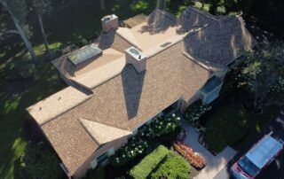 Aerial view of a house with brown shingled roof, chimney, and surrounding greenery and parked cars