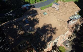 Aerial view of a tan-roofed house with skylights, trees casting shadows, and a driveway beside it
