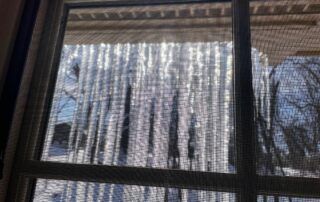 Snow-covered icicles hanging outside a window with bare trees in the background