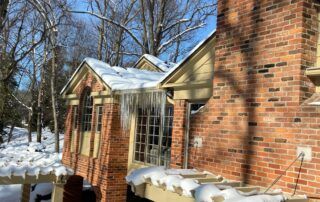 Snow-covered brick house with large windows and a blue sky behind leafless trees