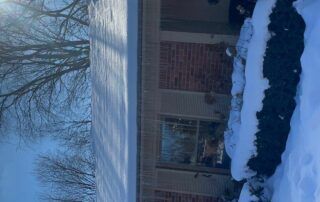 Snow-covered brick house with a front porch, shrubs, and bare trees in winter.