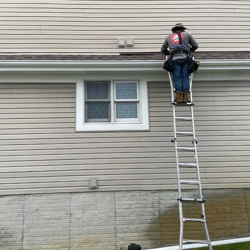 Worker on a ladder inspecting the side of a beige house near a window and roofline.