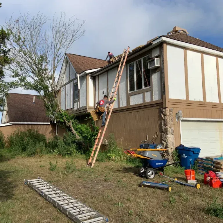 Worker on a ladder painting the exterior of a beige house beside a wheelbarrow and tools.