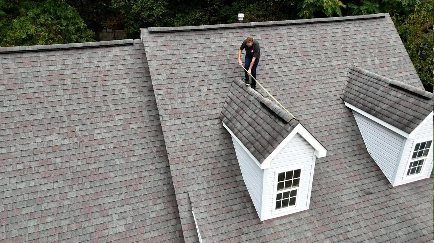 Worker on a gray shingled roof beside dormer windows, inspecting or repairing the roof.