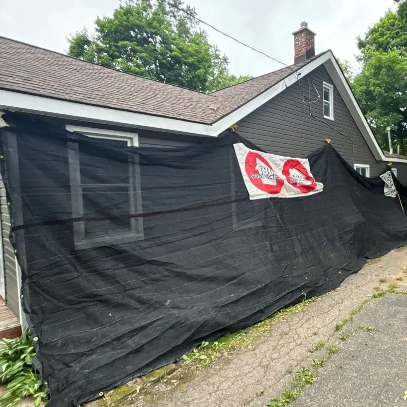 House wrapped in black tarp with a white and red sign on the side, beside a cracked driveway.