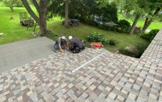 Two workers fixing a shingled roof beside a green yard and trees