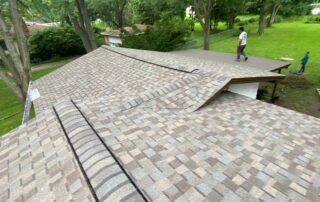 View of a shingled roof in a wooded area, with two people standing in the background.