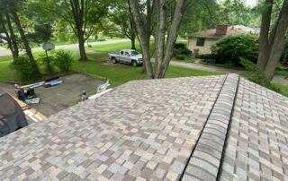 View from a shingled roof over a driveway, yard, and parked car beneath tall trees