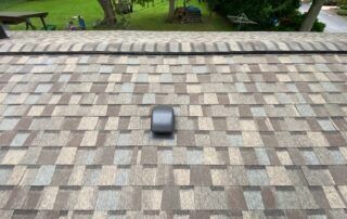 Small gray device on a shingled rooftop with grass and trees in the background