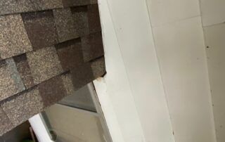 Close-up of a house roof with brown shingles beside a white wall and window frame.