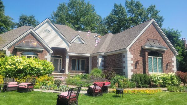 Brick suburban house with manicured lawn, flower beds, and outdoor chairs under a bright blue sky