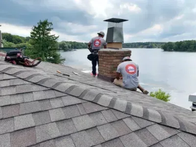 Two workers repairing a lakeside roof near a stone chimney and red ATV over the water