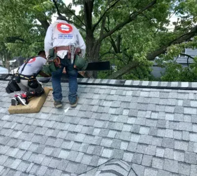 Two roofers repairing gray shingles on a steep roof, with trees in the background.