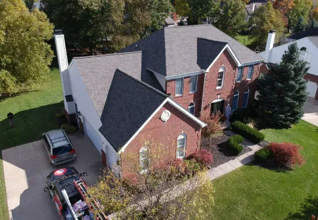 Aerial view of a red-brick house with a dark roof, driveway, parked cars, and green lawn.