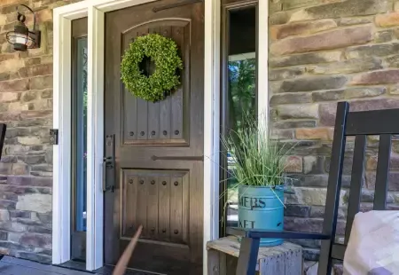 Front porch with stone walls, a wooden front door, wreath, and chair beside a blue planter.