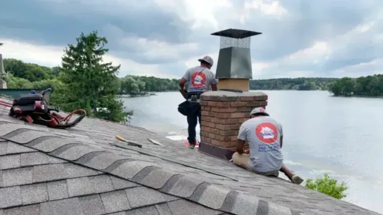 Two workers on a sloped roof repairing a brick chimney beside a lake