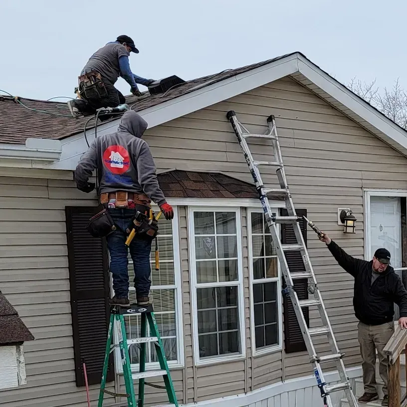 Workers repairing roof and siding on a house, with ladders set against the front wall.